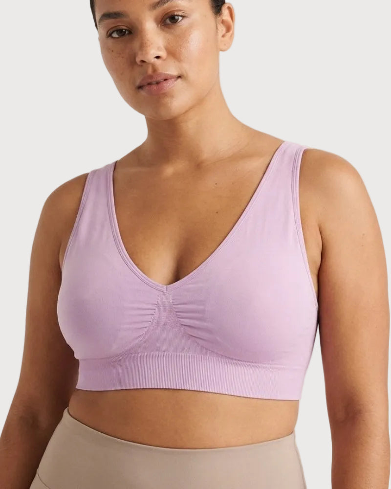 A woman wearing a light lavender bra with a deep V-neck and gathered center detail, paired with beige leggings, against a plain light background.